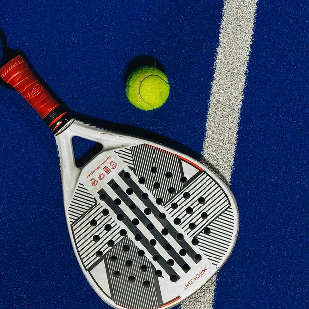 Nahaufnahme eines Padel-Schl&auml;gers und eines gelben Balls auf einem blauen Spielfeld mit wei&szlig;em Linienmuster.
