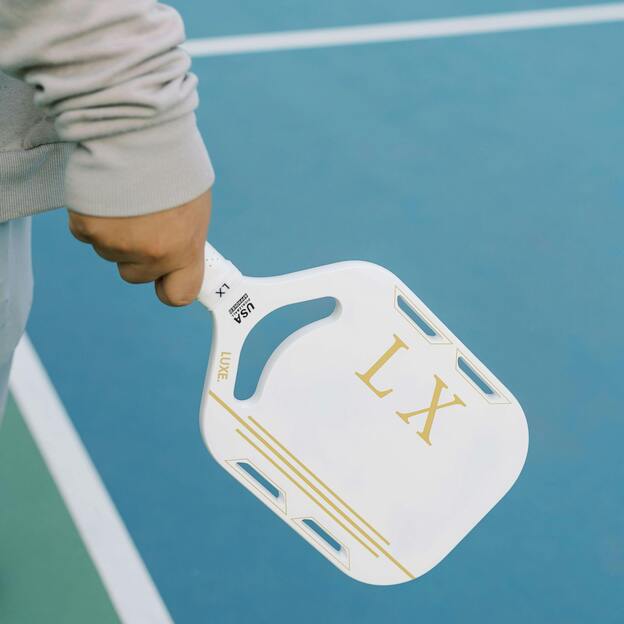 Nahaufnahme einer Person, die einen wei&szlig;en Paddel mit goldenen Schriftz&uuml;gen auf einer blauen Sportfl&auml;che h&auml;lt.
