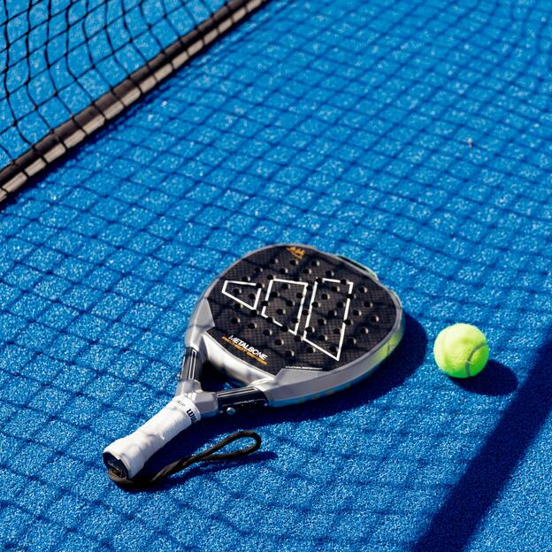 Nahaufnahme eines Padel-Schl&auml;gers und eines gelben Balls auf einem blauen Padelplatz neben dem Netz.