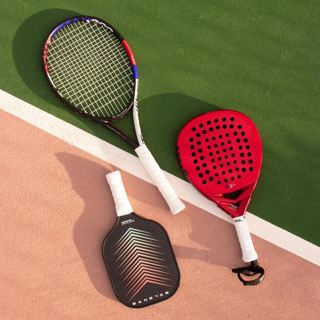 Drei Schl&auml;ger f&uuml;r verschiedene Sportarten liegen auf einem Tennisplatz: ein Tennisschl&auml;ger, ein Paddle-Schl&auml;ger und ein Pickleball-Schl&auml;ger.