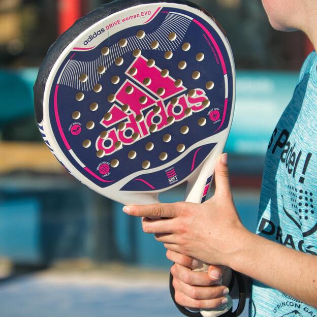 Nahaufnahme einer Person mit einem adidas Padel-Schl&auml;ger in der Hand, im Hintergrund eine Padel-Anlage.