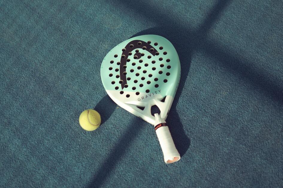 Nahaufnahme eines Padel-Schl&auml;gers mit gelbem Ball auf einer blauen Sportfl&auml;che.