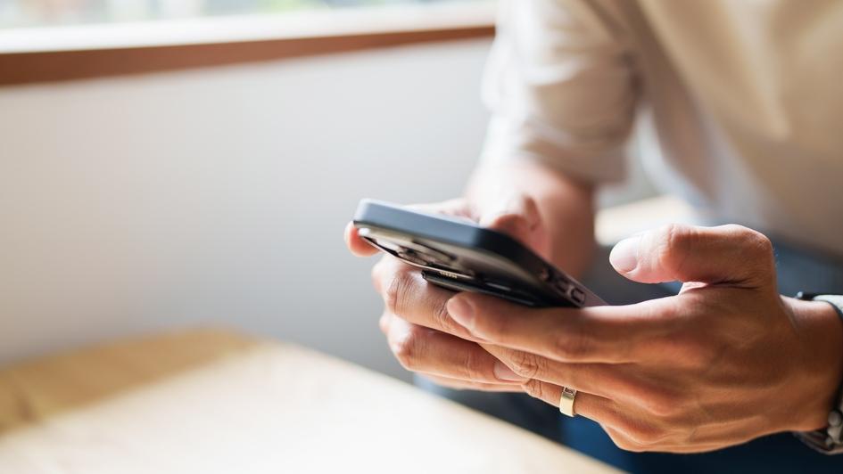 Nahaufnahme von H&auml;nden, die ein Smartphone halten, mit einem Tisch und Fenster im Hintergrund.