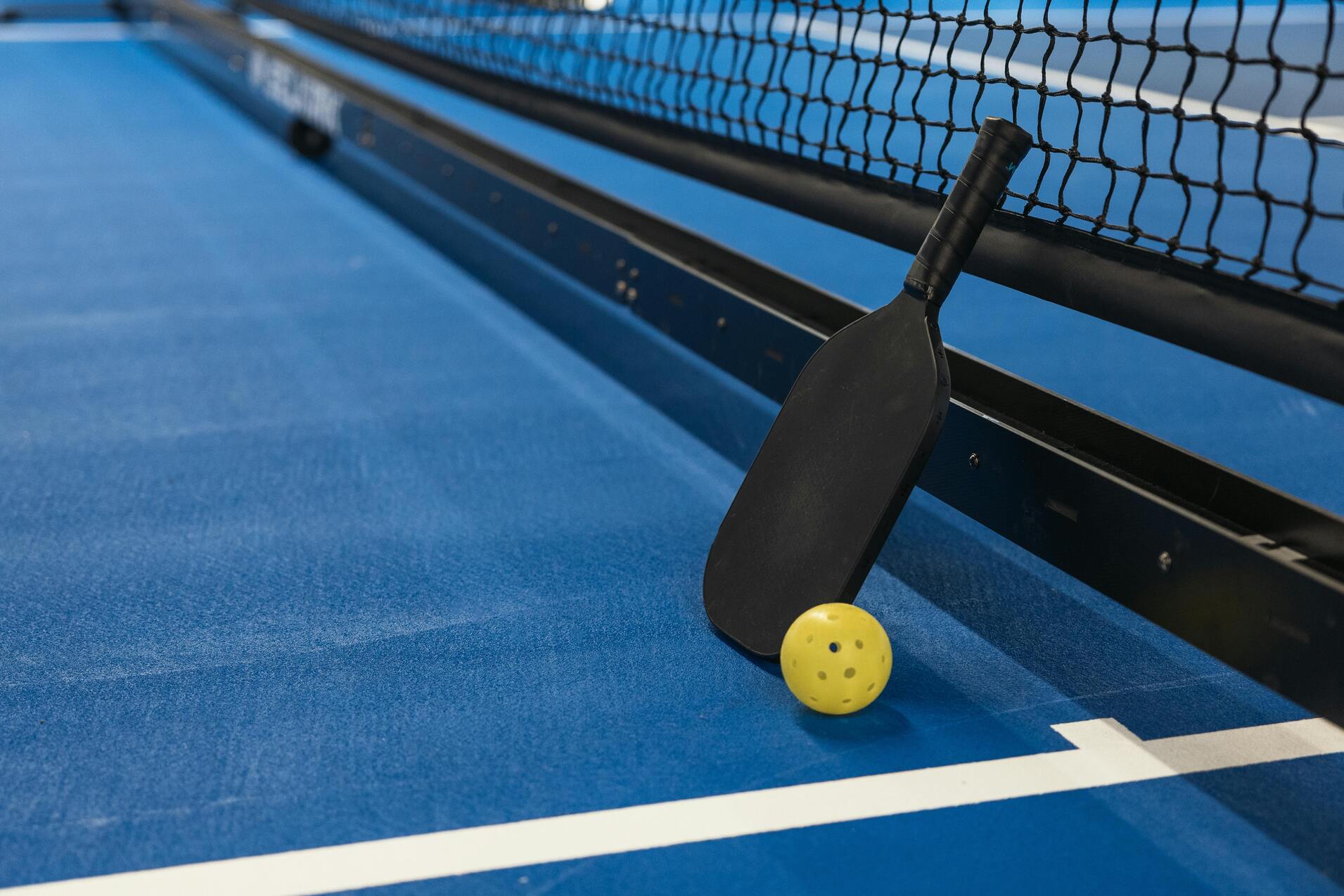 Nahaufnahme eines Paddles und eines gelben Balls neben einem Netz auf einem blauen Spielfeld.